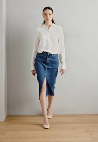 Bela srajca s gumbi, kombinirana z modrim jeans krilom z visokim pasom, ki ima sprednji razporek; model nosi bež petke in stoji na lesenem tleh.