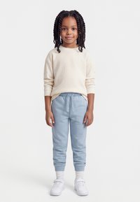 Νεαρή κοπέλα με πλεξούδες, φοράει κρεμ πουλόβερ, ανοιχτό μπλε παντελόνι φόρμας, λευκές κάλτσες και λευκά αθλητικά παπούτσια, στέκεται μπροστά σε απλό φόντο.