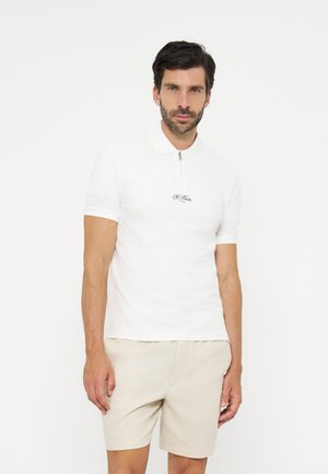 Homme aux cheveux courts et foncés avec une barbe, portant un polo blanc à manches courtes avec fermeture éclair et un short beige, debout devant un fond blanc.