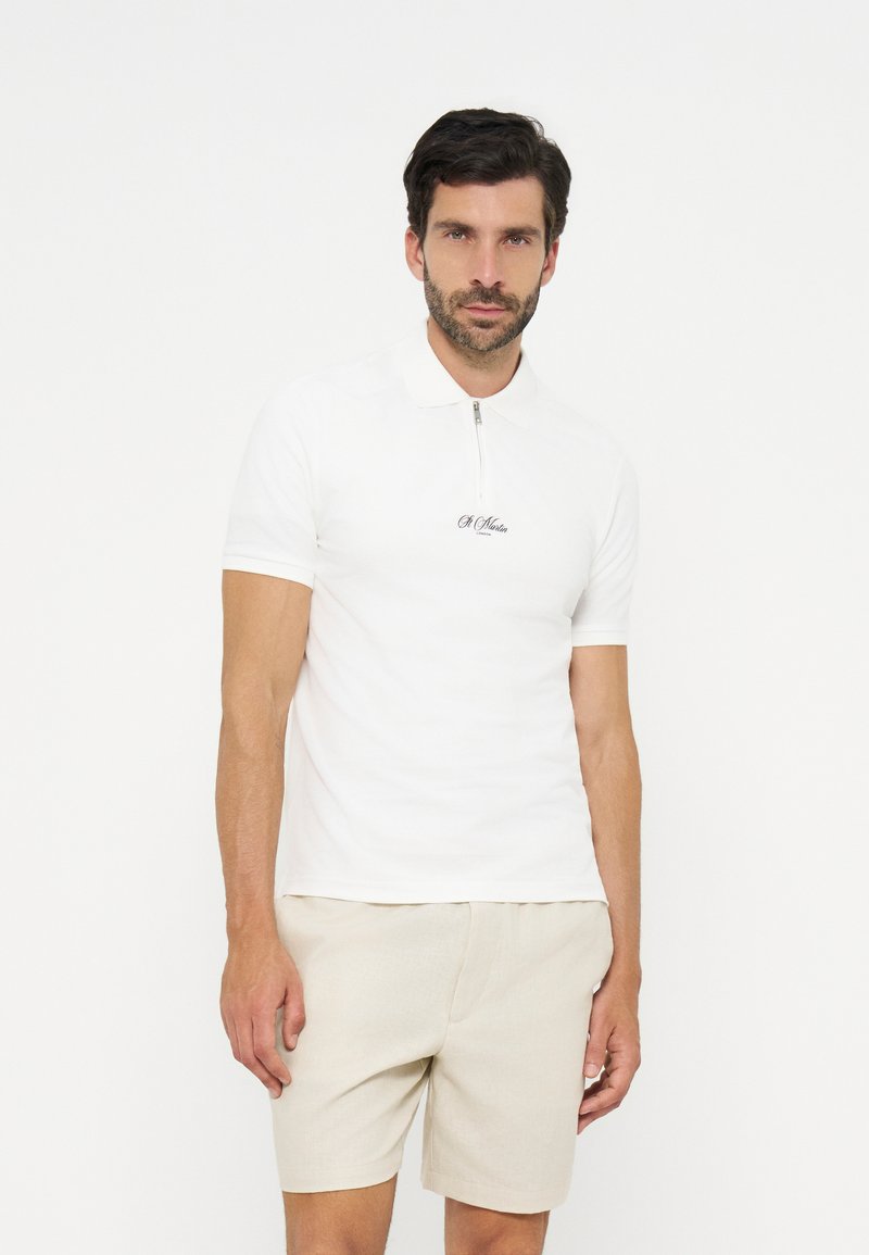 Homme aux cheveux courts et foncés avec une barbe, portant un polo blanc à manches courtes avec fermeture éclair et un short beige, debout devant un fond blanc.