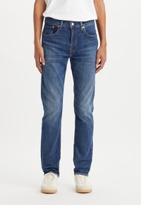 Personne portant un jean en denim bleu à jambes droites, des baskets blanches et un T-shirt blanc uni rentré, debout devant un fond blanc.