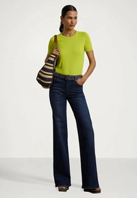 Mulher com uma blusa de manga curta verde-lima, calças de ganga escuras de cintura alta e pernas largas, mala de ombro tecida às riscas e sandálias de plataforma, em pé com uma mão no bolso.