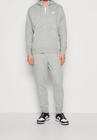 Grå zip-up hoodie och matchande sweatpants i mjukt material, med en liten vit logotyp på bröstet och ärmsluten. Svart och vit sneakers.