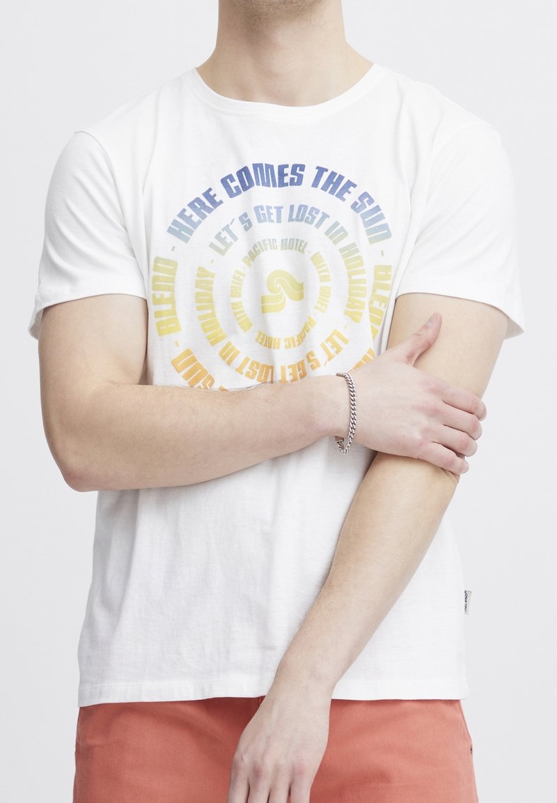 Homme portant un t-shirt blanc avec un texte circulaire coloré et un pantalon orange, tenant son bras gauche avec sa main droite.