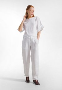 Valkoinen löysämallinen pellavapusero leveillä hihoilla ja siihen sopivat valkoiset pellavahousut, joissa on nyöritys ja etutaskut, yhdistettynä viininpunaisiin kenkiin.