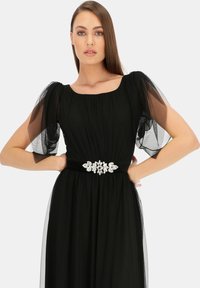 Czarna sukienka z przezroczystymi, bufiastymi rękawami, kwadratowym dekoltem i ozdobnym paskiem z biżuterią w talii; lekka, zwiewna tkanina.