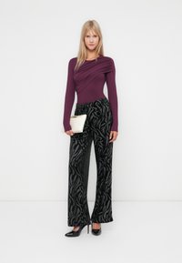 Haut mauve à manches longues avec un design drapé, associé à un pantalon noir à jambe large présentant un motif de feuille argentée, le tout accessoirisé avec une pochette blanche.