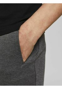 Graue Baumwollmischungshosen mit einer Nahaufnahme einer Hand in der Seitentasche, die die Textur des Stoffes und das Taschendesign zeigt. Schwarzes Oberteil sichtbar darüber.