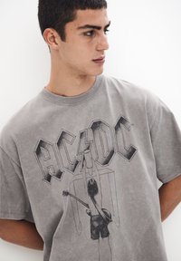 Tricou din bumbac gri cu mânecă scurtă, având un grafic negru cu un muzician ținând o chitară, textul "AC/DC" și un design vintage.