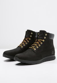 Stivali ankle in suede nero con occhielli e lacci dorati, con tacco in pelle nera e suola in gomma texturizzata. Logo Timberland sul lato.
