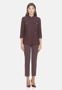 Blusa bordeaux scuro con colletto e due tasche sul petto, abbinata a pantaloni maroon su misura e tacchi alti lucidi bordeaux.