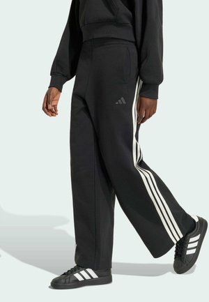 Osoba nosící černé teplákové kalhoty Adidas s bílými pruhy, která kráčí, doplněná o černé tenisky Adidas s bílými pruhy.
