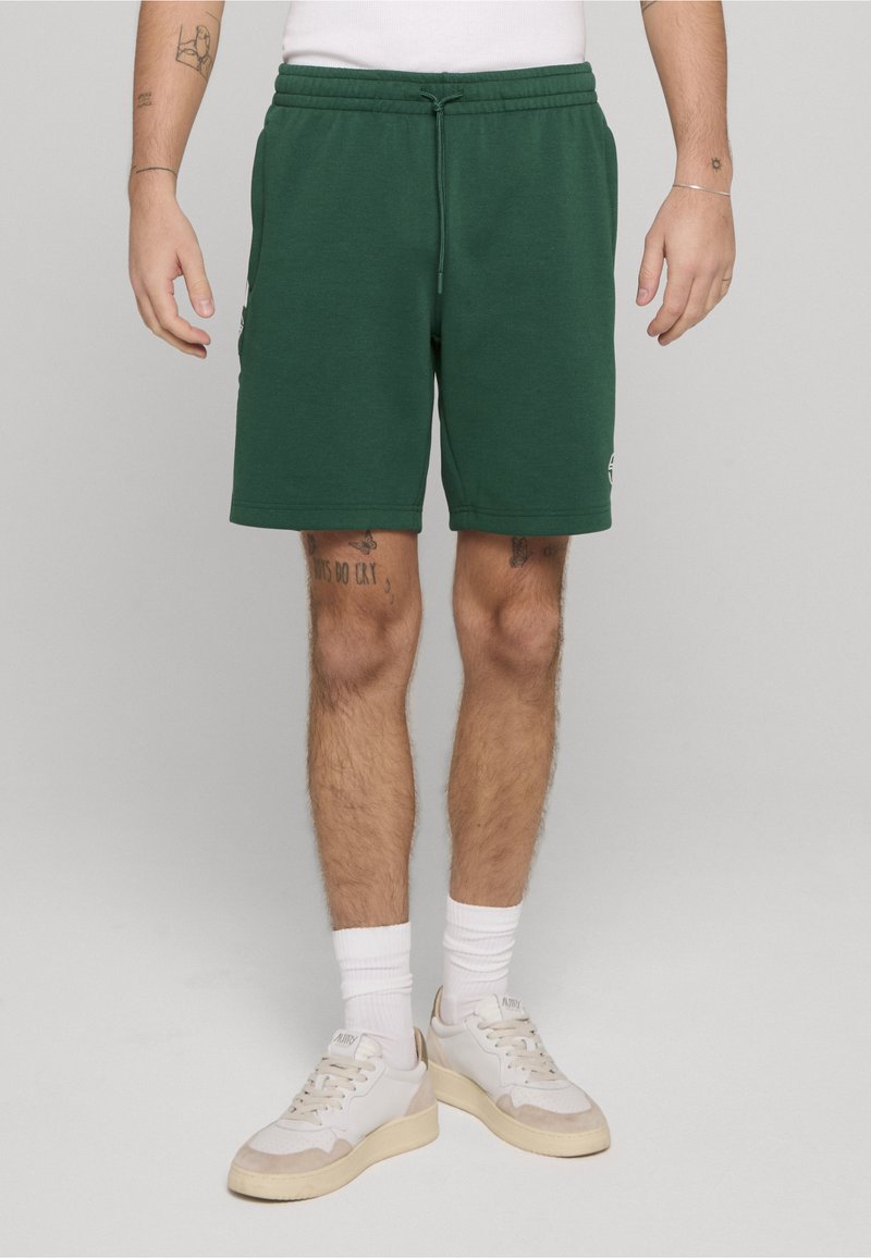 Groene shorts van een katoenmengsel met een elastische tailleband, trekkoorden, zijzakken en een klein logodetail aan de zoom. Gecombineerd met witte sneakers.