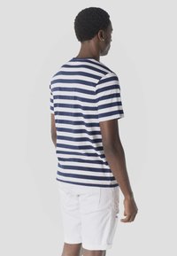 Homme de dos portant un t-shirt à manches courtes rayé marine et blanc et un short blanc, sur un fond uni gris clair.