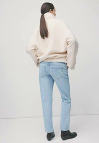 Kobieta z długimi ciemnymi włosami spiętymi w kucyk, ubrana w kremowy golf, jasnoniebieskie dżinsy i czarne botki, stojąca tyłem.