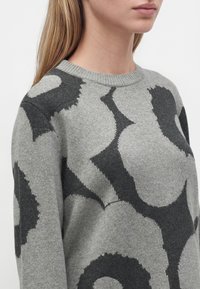 Gros plan d'une personne portant un pull en maille gris avec de grands motifs floraux foncés et un col rond côtelé.