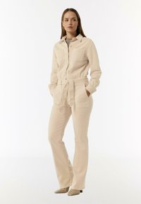 Jumpsuit beige à manches longues avec un col, fermeture à boutons, deux poches poitrine, taille cintrée et pantalon évasé ajusté.
