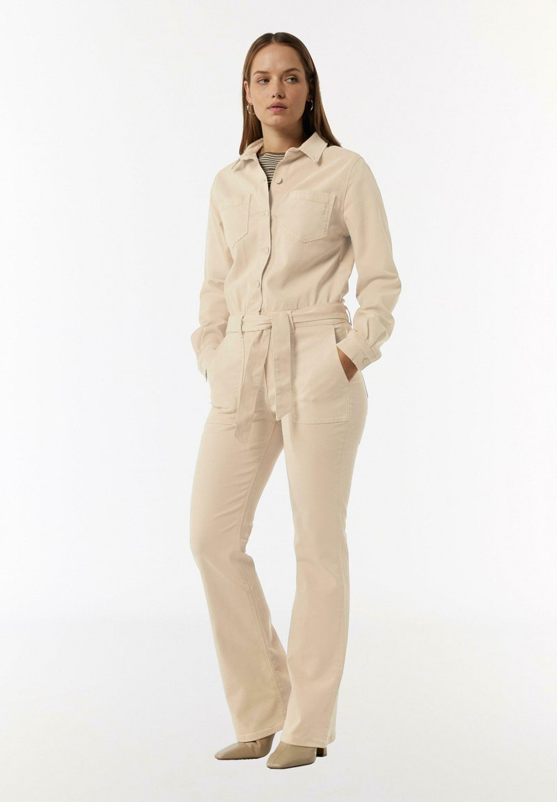 Jumpsuit beige à manches longues avec un col, fermeture à boutons, deux poches poitrine, taille cintrée et pantalon évasé ajusté.