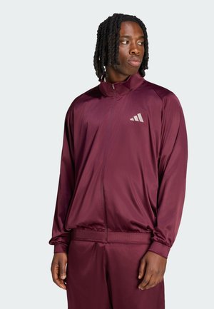 Homme aux cheveux tressés portant une veste et un pantalon de survêtement bordeaux, regardant sur le côté sur un fond clair uni.