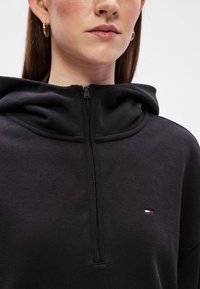 Μαύρο φούτερ με φερμουάρ και υψηλό γιακά, με ριμπ ραφές. Έχει ένα μικρό λογότυπο σε κόκκινο, λευκό και μπλε χρώμα στο στήθος. Μαλακή υφή.