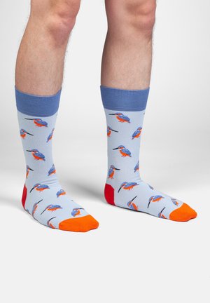 Männliche Unterschenkel, die hellblaue Socken mit blauen Bündchen, orangen Zehen, roten Fersen und einem wiederkehrenden Muster aus blauen und orangen Eisvögeln tragen.