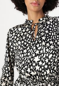 Blouse noir et blanc avec des motifs d'étoiles, dotée d'un col froncé, d'une ouverture en forme de trou de serrure et d'une taille élastique. Texture de tissu lisse.