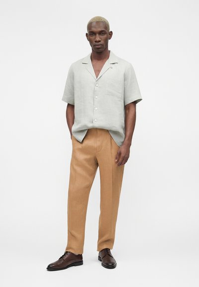 Άνδρας με κοντά ξανθά μαλλιά φοράει ανοιχτό γκρι πουκάμισο με κοντά μανίκια και κουμπιά, μπεζ παντελόνι και σκούρα καφέ επίσημα παπούτσια, στέκεται με τα χέρια στις τσέπες.