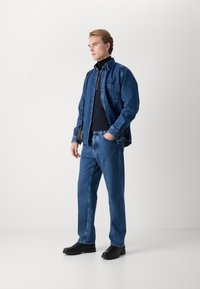 Camisa de denim superpuesta sobre un cuello de tortuga negro, combinada con jeans de denim azul y zapatillas negras sin cordones. El modelo está de pie en un fondo neutro.