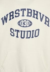 Krémová mikina s kapucí a tmavě modrým potiskem "WRSTBHVR" a "STUDIO." Má přední kapsu, měkkou strukturu a volný design.