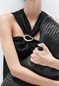 Borsa nera intrecciata con superficie testurizzata, tenuta contro un top in tessuto nero che presenta un prominente accento a forma di anello dorato sul collo.