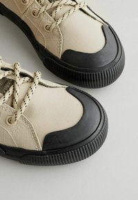 Beige Sneaker verfügen über ein Obermaterial aus strukturiertem Stoff mit einer schwarzen Gummizehenspitze und einer strukturierten Sohle. Schwarze und cremefarbene Schnürsenkel runden das Design ab.