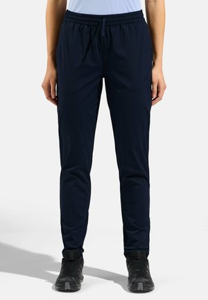 Personne portant un pantalon de sport fitted bleu marine foncé avec une ceinture élastique et des chaussures de randonnée noires, debout devant un fond uni.