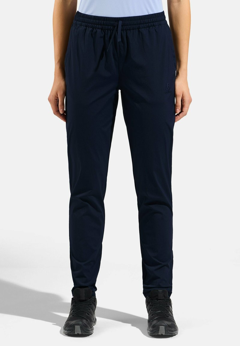 Personne portant un pantalon de sport fitted bleu marine foncé avec une ceinture élastique et des chaussures de randonnée noires, debout devant un fond uni.