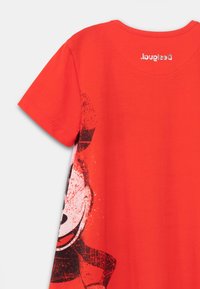 Czerwona bawełniana koszulka z grafiką przedstawiającą postać w czerni i bieli, z akcentami w postaci plam z jednej strony. Krótkie rękawy i okrągły dekolt.