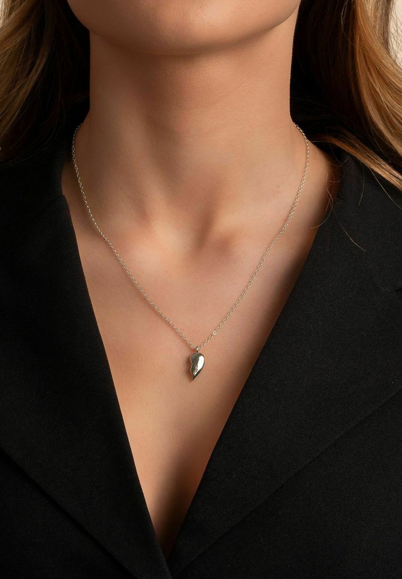 Silverfärgad kedjehalsband med en tårformad hänge med en slät yta, uppvisad mot ett svart plagg.