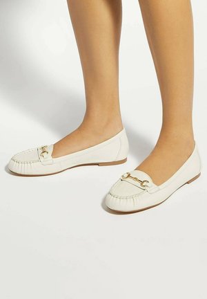 Jambes de femme portant des mocassins en cuir blanc avec un détail mors en or, debout sur un sol blanc uni.