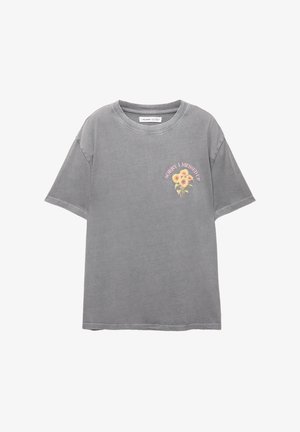 Siva oversized majica od pamuka, sa okruglim izrezom i kratkim rukavima. Ispred se nalazi print s ružičastim tekstom i ilustracijom suncokreta.