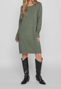 Robe-pull en maille vert olive avec manches longues et poignets côtelés, associée à des bottes en cuir noir montantes jusqu'aux genoux, dotées d'un bout pointu et d'une fermeture éclair.