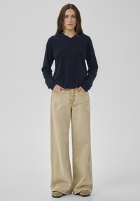 Marineblauer Pullover mit V-Ausschnitt, kombiniert mit beigen, weit geschnittenen Jeans. Pullover aus weichem Stoff, Jeans mit seitlichen Taschen und entspannter Passform.