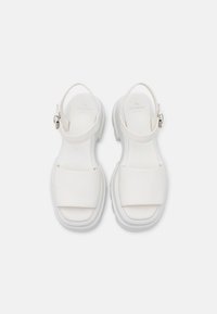 White leather sandals with a flat sole, wide front straps, and adjustable ankle buckle. Smooth texture with minimalistic design details.
