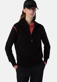 Felpa nera con zip, colletto e polsini a coste, con dettagli rossi sulle spalle e un tessuto strutturato. Indossata con un berretto nero.