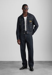 Jeansjakke og bukser sæt i mørk vask, med gul broderi og sølv hardware. Modellen bærer sorte solbriller og en hvid skjorte.