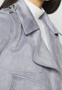 Zbliżenie na osobę wearing light-gray suede jacket with large lapels, shoulder epaulet, and a white high-neck top underneath.