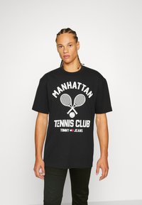 Juodas medvilnės marškinėliai su baltais spaudiniais, kuriuose yra užrašas "Manhattan Tennis Club" ir teniso raketės grafika. Laisvas kirpimas, apvalios iškirptės dizainas.
