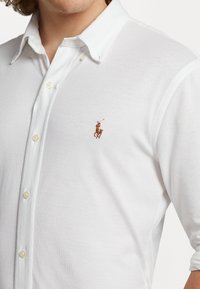 Vit långärmad polo skjorta i texturerat tyg, med en platt knappslå med tre knappar och en liten färgad broderad logotyp på vänster bröst.