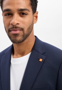 Mann mit kurzen lockigen Haaren und Bart, der einen marineblauen Blazer mit einem orangefarbenen "MOVE"-Anstecker und einem weißen Hemd vor weißem Hintergrund trägt.