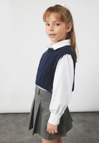 Mørkeblå strikket vest med fletmønster over en hvid skjorte med krave, parret med en grå plisseret nederdel, der viser et lagdelt outfitdesign.