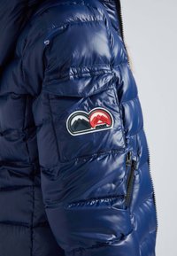 Manche de veste matelassée bleu marine avec poche zippée et écusson représentant des montagnes superposées en rouge et noir avec le texte "JACKIT".