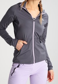 Mujer vistiendo una chaqueta gris ajustada con cremallera y agujeros para los pulgares, y pantalones de color púrpura claro con un bolsillo con cremallera lateral, de pie con una mano en el bolsillo.