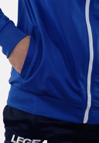 Blauwe sportjack met witte rits, met zijzakken en een gladde textuur. Gecombineerd met marineblauwe shorts met 'LEGEA' merknaam.